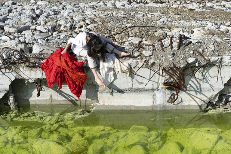 Dancer Eiko Otate sits on a wall next to a still body of water.